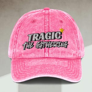 May include: A pink, distressed baseball cap with the text "TRAGIC THE GATHERING" embroidered in white and black. A yellow star is embroidered above the word "GATHERING". The cap has a curved brim and a pink button on top.