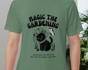 May include: Sage green t-shirt with the text "MAGIC THE GARDENING" in a bold, stylized font. The graphic features a black cat wearing a mushroom hat, a butterfly, and mushroom illustrations. The text "WHEREVER I SET MY PACK AND REST MY HEAD, I AM HOME" is below the graphic.