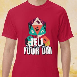 Puede incluir: Camiseta roja con un gráfico de dados y el texto "TELL You love them YOUR DM". Los dados son de varios colores, incluyendo verde azulado, naranja y morado. La camiseta es de cuello redondo y manga corta.