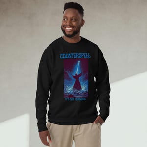 Peut inclure: Sweat-shirt noir avec le mot "COUNTERSPELL" en bleu au-dessus d'un dessin d'une silhouette les bras levés, éclairée par un rayon de lumière. Le texte "IT'S NOT PERSONAL" est en dessous. Le sweat-shirt est porté par une personne.