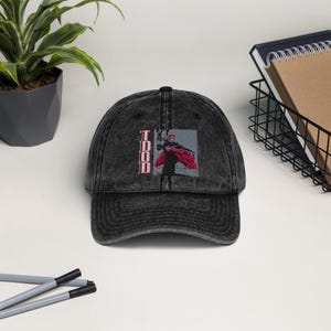 May include: A black, distressed baseball cap with a graphic of a figure in red and gray. The word "TODD" is vertically printed in red on the left side of the cap. The cap is sitting on a white surface.
