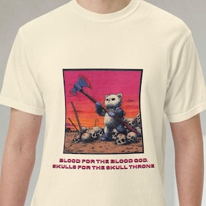 以下が含まれることがあります： クリーム色のTシャツ。斧を持った猫と頭蓋骨のグラフィック、テキスト「BLOOD FOR THE BLOOD GOD, SKULLS FOR THE SKULL THRONE」が描かれています。デザインはピンクとオレンジの背景です。
