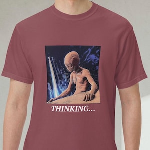 May include: Maroon t-shirt featuring a graphic of an alien figure with a cityscape background. The alien is depicted in a contemplative pose, with the text "THINKING..." below the image. The shirt is made of a soft material.
