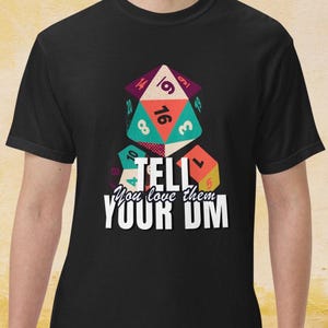 May include: Black t-shirt featuring a colorful graphic of dice with numbers and the text "TELL You love them YOUR DM" in white. The dice are in various colors, including purple, teal, orange, and yellow. The shirt is a crew neck.