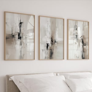 Può includere: Un set di tre stampe artistiche da parete rettangolari verticali in cornici di legno chiaro. Ogni stampa presenta una composizione astratta con toni neutri beige, bianco e nero, caratterizzata da pennellate verticali e sottili linee orizzontali.