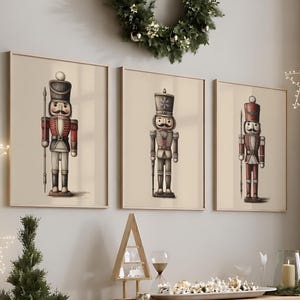 Puede incluir: Tres cuadros de pared con ilustraciones de soldados cascanueces en uniformes de color rojo, blanco y gris. Cada lámina muestra a un soldado de pie con un sombrero alto y un bastón, presentados en un marco sencillo de color claro.