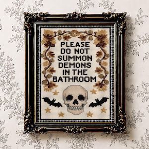 May include: A framed cross-stitch embroidery featuring the black text PLEASE DO NOT SUMMON DEMONS IN THE BATHROOM, surrounded by brown autumn leaves, small gold stars, two black bats, and a central beige skull in a dark ornate frame.