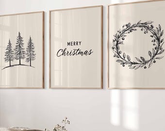 May include: Three framed art prints with a neutral background. One print features three stylized pine trees, another says "MERRY Christmas" in script, and the third shows a wreath. The frames are a light wood color.