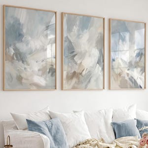May include: Three rectangular wall art prints in light wooden frames featuring abstract brushstrokes in shades of blue, white, and beige. The artwork displays layered, sweeping paint textures across each vertical canvas, creating a cohesive set.