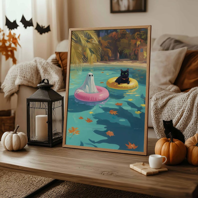 May include: A framed art print depicts a ghost in a pink inflatable ring and a black cat in a yellow ring floating in a pool. The artwork has a wooden frame and is displayed on a wooden table with pumpkins and a lantern.