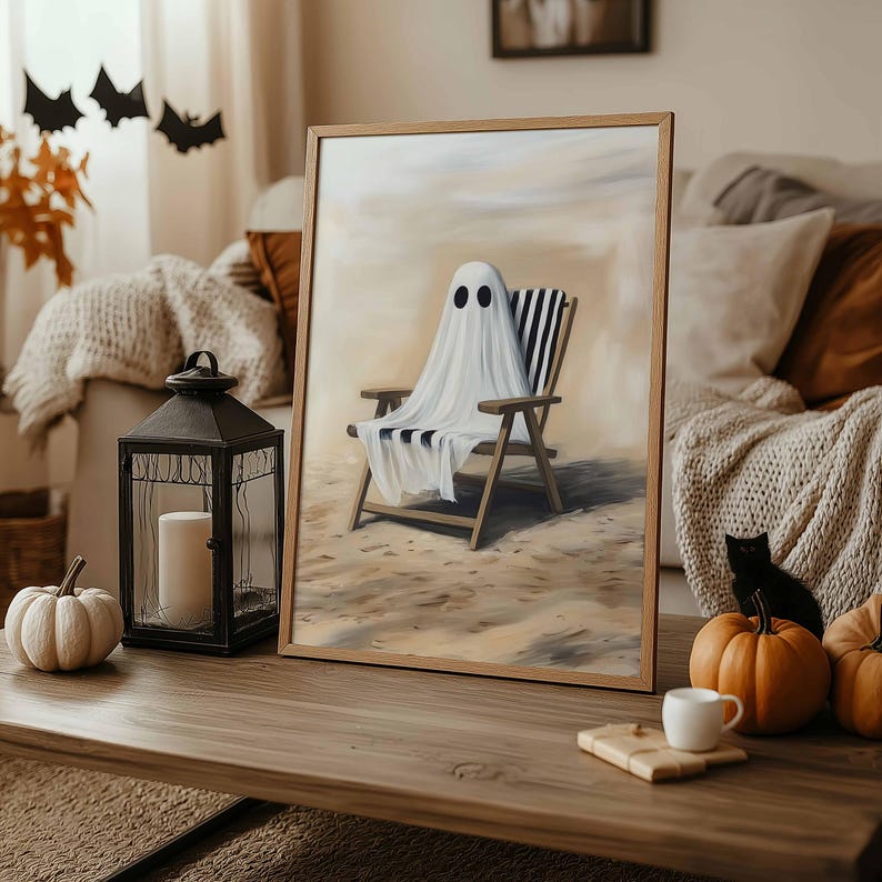 K&ouml;nnte beinhalten: Ein gerahmtes Kunstwerk zeigt einen Geist, der &uuml;ber einem Strandstuhl mit schwarz-wei&szlig;en Streifen drapiert ist. Der Geist hat zwei schwarze Augen. Das Kunstwerk steht auf einem Holztisch mit einer Laterne, K&uuml;rbissen und einer schwarzen Katze, was ein Halloween-Thema ergibt.