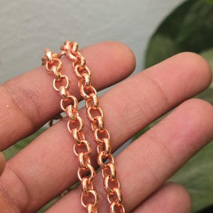 Puede incluir: Primer plano de un collar de cadena de oro rosa. La cadena está hecha de eslabones ovalados entrelazados. El collar se muestra sobre un fondo borroso.