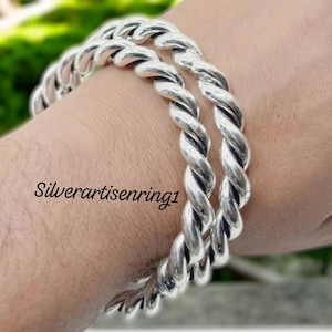 May include: Two silver-coloured, twisted-rope style bracelets. The bracelets are worn on a person's wrist. The image has the text "Silverartisenring1" in the lower left corner. The bracelets are made of a shiny metal.