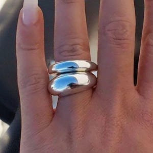 Könnte beinhalten: Zwei glänzende, silberne Ringe, die auf einem Finger gestapelt sind. Die Ringe haben ein glattes, abgerundetes Design, das das Licht reflektiert. Ein schlichtes, elegantes Schmuckstück.