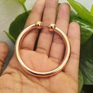 Puede incluir: Una pulsera de brazalete abierta de color cobre, con un diseño liso y redondeado. La pulsera presenta una bola esférica en cada extremo. La joya se sostiene en una mano, con follaje verde en el fondo.