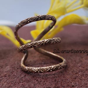 May include: A bronze-colored ring with an intricate geometric pattern. The ring has a unique, open design that wraps around the finger. The ring is displayed against a blurred background with yellow flowers and a brown surface.