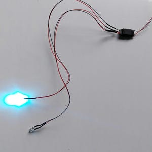 May include: A close-up of a circuit board with wires connected to a small, round, blue LED light. The LED is lit and glowing brightly.