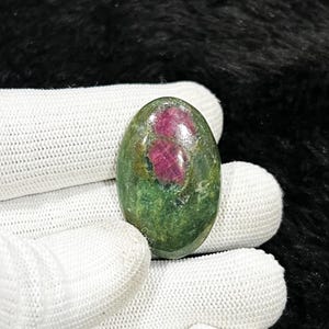 May include: An oval gemstone with a green base and a striking red inclusion. The polished stone has a smooth surface and a natural appearance. The gemstone is held by a gloved hand.