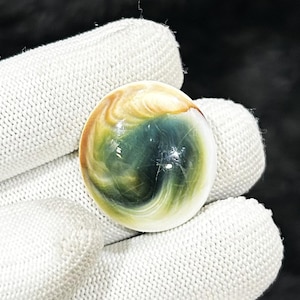May include: A polished, spherical gemstone with swirling patterns of green, yellow, and cream colors. The stone is held in a white glove, showcasing its smooth, reflective surface. The gemstone's design is reminiscent of a miniature planet.