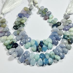 May include: Three strands of gemstone beads in shades of blue, green, and purple. The teardrop-shaped beads are strung together, with white tassel accents at the top. The beads are arranged on a white surface.