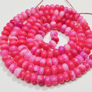 May include: A coiled strand of pink and magenta beads. The beads are oval-shaped and strung on a thin, reddish-brown string. The beads vary in shade, creating a gradient effect. The background is a plain white surface.