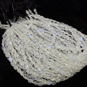 May include: A collection of faceted, translucent white moonstone beads strung on multiple black threads. Each small, rectangular-shaped bead displays a subtle, iridescent blue adularescence, creating a shimmering effect across the entire cluster of gemstone strands.