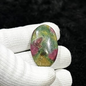 May include: An oval-shaped gemstone with a mix of colors. The stone features a base of green and yellow with patches of deep red. The gemstone is held in a white glove against a black background.