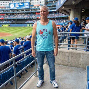 May include: A light blue tank top with the text "JAYS GO" and a blue bird logo. The person is wearing blue jeans and white sneakers. The background shows a baseball stadium filled with fans.