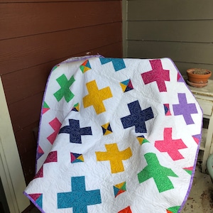 May include: A white quilt with a purple border and a patchwork design featuring colorful plus signs. The plus signs are in various colors, including red, orange, yellow, green, blue, and purple.
