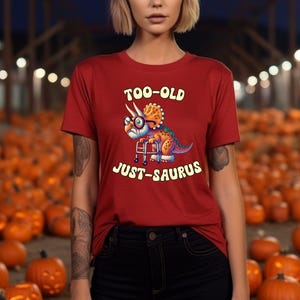 Puede incluir: Camiseta roja con el texto "TOO-OLD JUST-SAURUS" y un dinosaurio de dibujos animados con gafas y andador. La camiseta está hecha de un material suave.