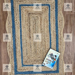 May include: A rectangular jute rug with a wavy border and a blue border. The rug has a natural, woven texture. A book with the text "INDIA CARPET EXPO 47" is on the rug. The rug is on a wooden surface.
