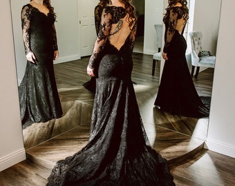May include: A woman in a black lace wedding dress with a low back and long sleeves stands in front of a mirror. The dress has a fitted bodice and a flowing skirt. The woman is wearing her hair down and has a flower in her hair.