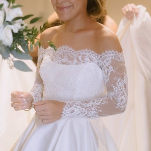 May include: A white off-the-shoulder wedding dress with long lace sleeves. The bride is smiling, holding a bouquet of white flowers. The dress has a satin skirt and a lace bodice.