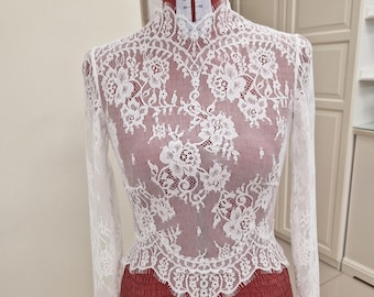 May include: A white lace top with long sleeves and a high neckline. The intricate floral pattern of the lace is visible. The top is displayed on a red dress form.
