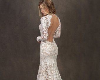 May include: A white lace wedding dress with a low back and long sleeves. The dress has a fitted bodice and a flowing skirt.