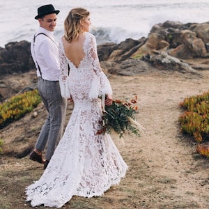 May include: A bride and groom stand on a rocky path overlooking the ocean. The bride is wearing a white lace wedding dress with long sleeves and a low back. The groom is wearing a white shirt, grey pants, and a black hat. They are both looking out at the ocean.