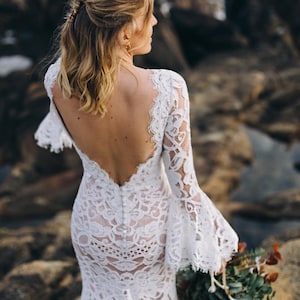 May include: A floor-length white lace dress featuring a deep open back, long bell sleeves with scalloped edges, and a vertical row of small white buttons running down the center back. The intricate floral lace pattern overlays a neutral-toned lining.
