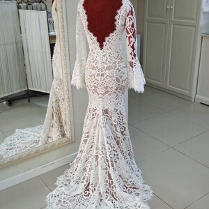 May include: A floor-length white lace gown featuring long bell sleeves, a deep scoop back, and a row of small white buttons running down the center back. The dress has a fitted bodice that flares into a train, showcasing intricate floral lace patterns.