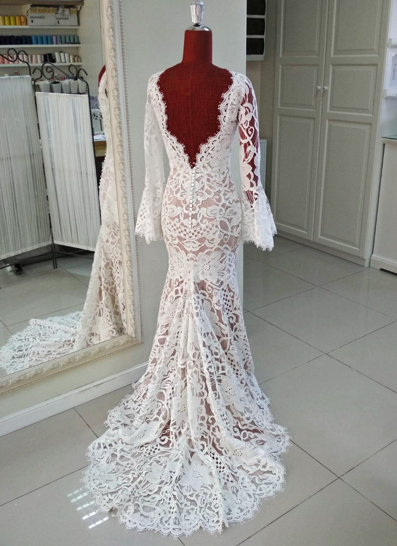 May include: A floor-length white lace gown featuring long bell sleeves, a deep scoop back, and a row of small white buttons running down the center back. The dress has a fitted bodice that flares into a train, showcasing intricate floral lace patterns.