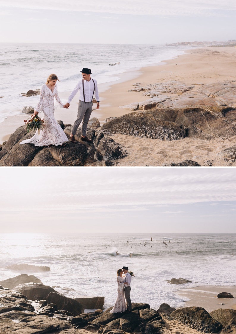 May include: A couple standing on coastal rocks by the ocean. One individual wears a long-sleeved white lace gown and holds a floral bouquet. The other wears a white button-down shirt, grey trousers, brown suspenders, and a dark fedora hat.