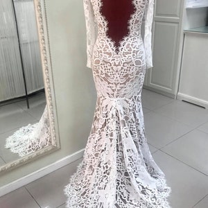 May include: A floor-length white lace gown with long sleeves and a deep, open back design. The dress features intricate floral lace patterns throughout, a fitted bodice, and a flared skirt that extends into a small train at the floor.