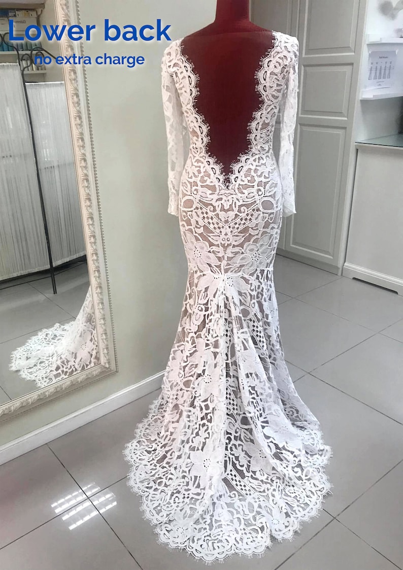 May include: A floor-length white lace gown with long sleeves and a deep, open back design. The dress features intricate floral lace patterns throughout, a fitted bodice, and a flared skirt that extends into a small train at the floor.
