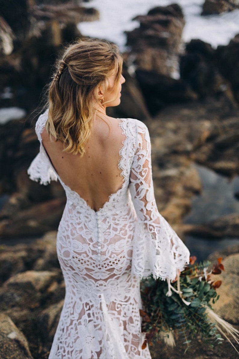 May include: A floor-length white lace dress featuring a deep open back, long bell sleeves with scalloped edges, and a vertical row of small white buttons running down the center back. The intricate floral lace pattern overlays a neutral-toned lining.