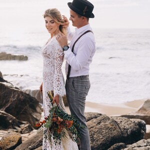 May include: A white lace long-sleeve dress with a V-neckline, a white button-down shirt, brown suspenders, gray trousers, a black fedora with a small feather detail, and a bouquet of dried eucalyptus and pampas grass on a rocky beach.