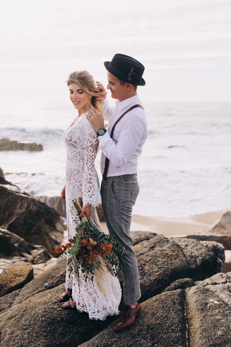 May include: A white lace long-sleeve dress with a V-neckline, a white button-down shirt, brown suspenders, gray trousers, a black fedora with a small feather detail, and a bouquet of dried eucalyptus and pampas grass on a rocky beach.