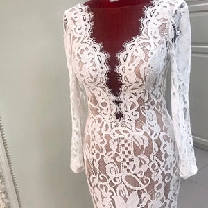 May include: A white long-sleeved lace dress with a deep V-neckline and a nude-colored lining. The dress features intricate floral lace patterns and scalloped edges along the neckline. Text on the image reads: Lower neckline no extra charge.