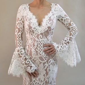 May include: A floor-length white lace dress featuring long, flared bell sleeves and a deep V-neckline. The garment is constructed from intricate floral lace patterns with scalloped edges along the neckline and sleeve cuffs, creating a textured look.