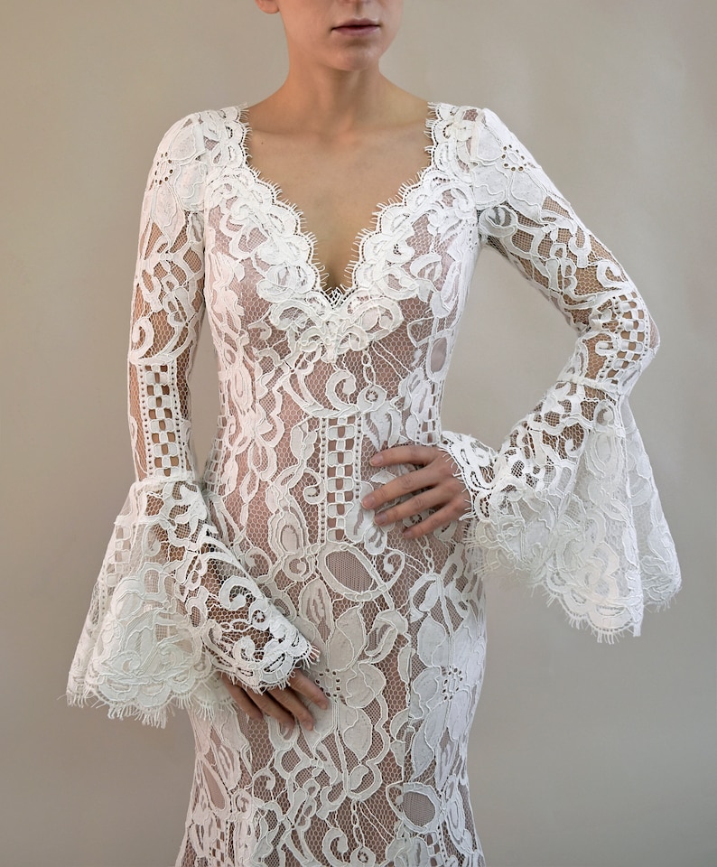 May include: A floor-length white lace dress featuring long, flared bell sleeves and a deep V-neckline. The garment is constructed from intricate floral lace patterns with scalloped edges along the neckline and sleeve cuffs, creating a textured look.