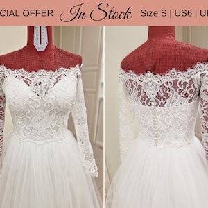 May include: White off-the-shoulder wedding dress with long lace sleeves and a tulle skirt. The dress is on a mannequin and has a button closure in the back. The text "In Stock" and "Size S | US6 | UK10" is visible in the image.