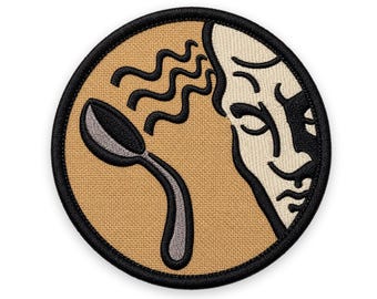 May include: A circular embroidered patch with a tan background and a thick black border. The design features a stylized human face profile on the right side and a silver spoon on the left, with black wavy lines emanating from the spoon.